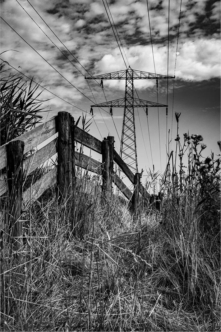 Een verweerd houten hek en wild grasland vormen de voorgrond van dit contrastrijke landschap. Daarboven rijst een imposante hoogspanningsmast op tegen een dramatische wolkenlucht. De grillige natuur en strakke lijnen van de elektriciteitsdraden creëren een intrigerend samenspel tussen rust, ruigheid en menselijke infrastructuur.