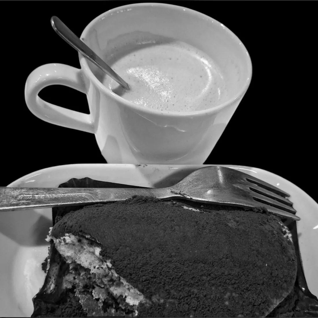 Zwart-witbeeld van een kopje koffie met melkschuim en een lepel erin, boven een bord met een stuk dessert met cacaopoeder en een vork, tegen een egaal zwarte achtergrond, met weerspiegelend licht op het witte porselein en het metalen bestek.