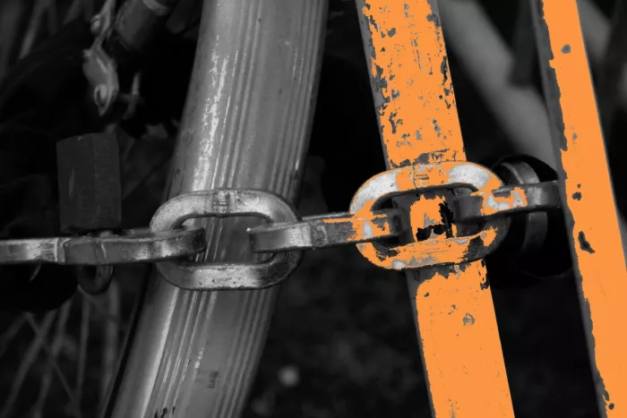 Close-up van een zware ketting door een fietsframe, in zwart-wit met oranje, afgebladderde verf op de buizen.