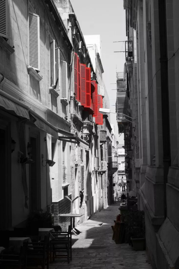 Smalle straat in Malta tussen kalkstenen gevels in zwart-wit, waar enkel de luiken van een houten balkon rood zijn.