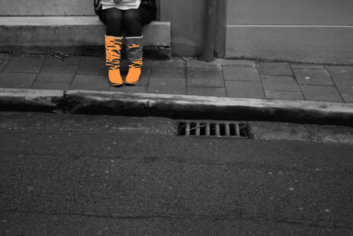Zittende persoon met oranje laarzen op een stoeprand, boven een rioolrooster, in verder zwart-wit straatbeeld.