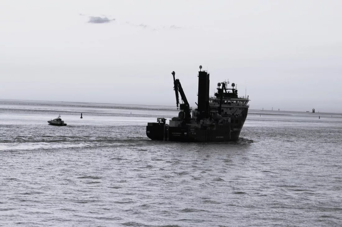 Werkschip met kranen en een kleine boot op rimpelend water onder een bleke, witte lucht