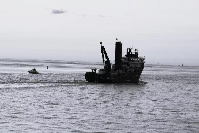 Werkschip met kranen en een kleine boot op rimpelend water onder een bleke, witte lucht