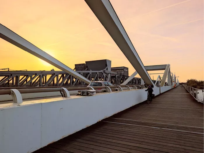Witte stalen brugbogen boven een houten fietspad op de Temsebrug, met een fietser en een auto bij zonsopgang