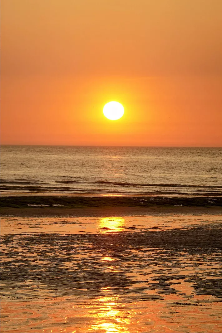 Grote zon net boven de zee in een oranje lucht, met een gouden lichtbaan op het natte strand