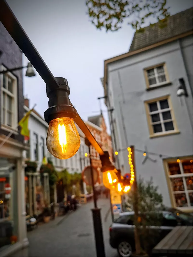 Grote gloeilamp aan een paal in een straat met witte gevels en lichtsnoeren