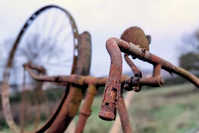 Roestig fietsstuur met een aangevreten handvat en een overgroeide bel staat scherp in beeld, met een onscherp wiel en een groen weiland met bomen op de achtergrond onder een bleke lucht.