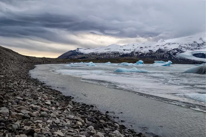 Gletsjermeer met drijvend ijs, een keienoever en besneeuwde bergen onder grijze wolken