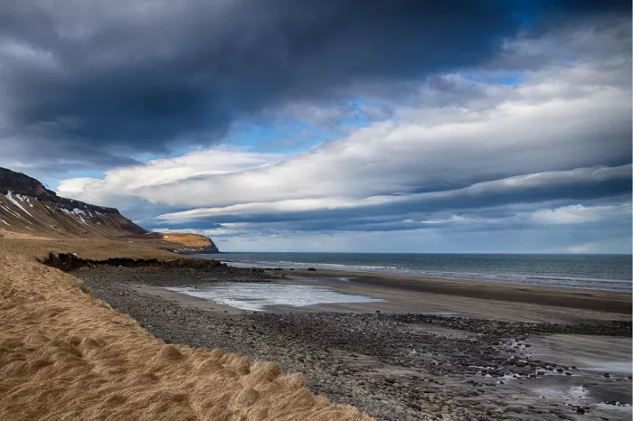 Kiezelstrand met goudgeel gras en donkere dreigende wolken boven de zee