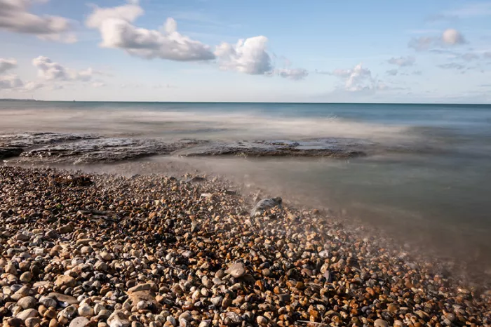 Kiezelstrand met platte rotsen in een nevelige zee onder een blauwe lucht met witte wolken