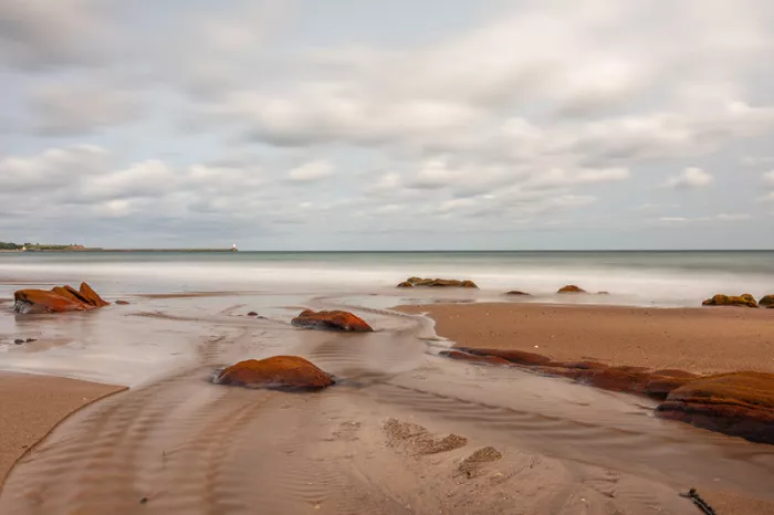 Zandstrand met roodbruine rotsen en zacht stromend zeewater onder een bewolkte lucht