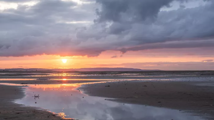 Zonsondergang boven een strand bij eb met zware grijze wolken en een plas die de oranje gloed spiegelt