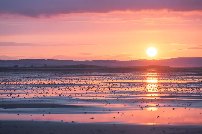 Ondergaande zon boven heuvels met een roze lucht en gouden reflecties op een nat strand met stenen