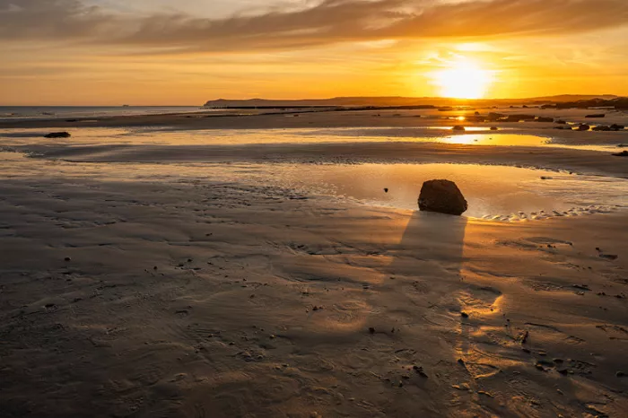 Lage zon boven een strand bij eb, met een losse rots die een lange schaduw werpt over het zand