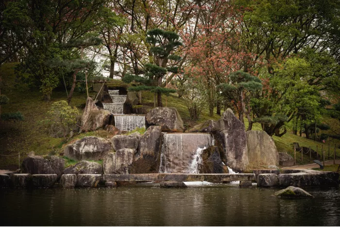 Trapsgewijze waterval tussen grote rotsblokken en gesnoeide dennen, die uitkomt in een stille vijver in de Japanse tuin van Hasselt.