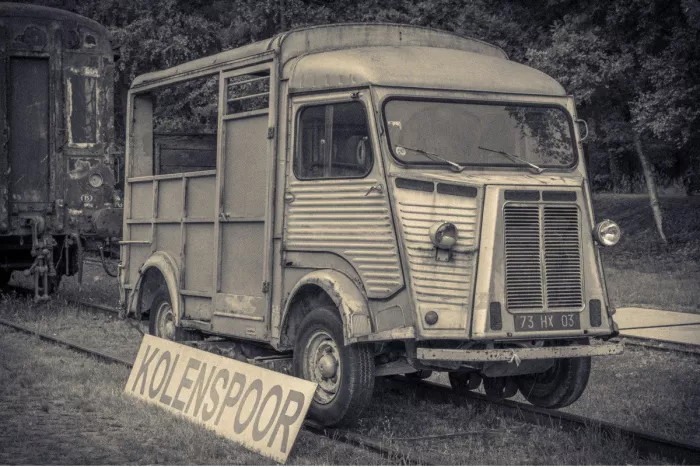 Verweerde oude Citroën-bestelwagen op de sporen bij het oude stationnetje van As, met een bord Kolenspoor en een roestige treinwagon ernaast.