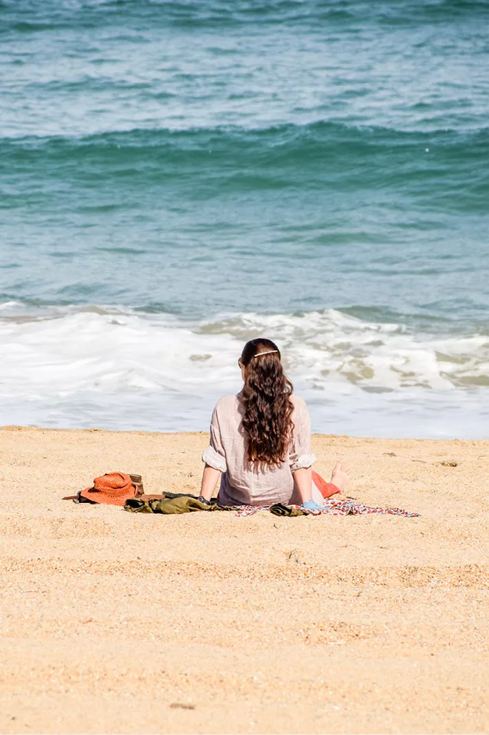 Vrouw met lang krullend haar zit op een dekentje op het strand en kijkt naar de golven