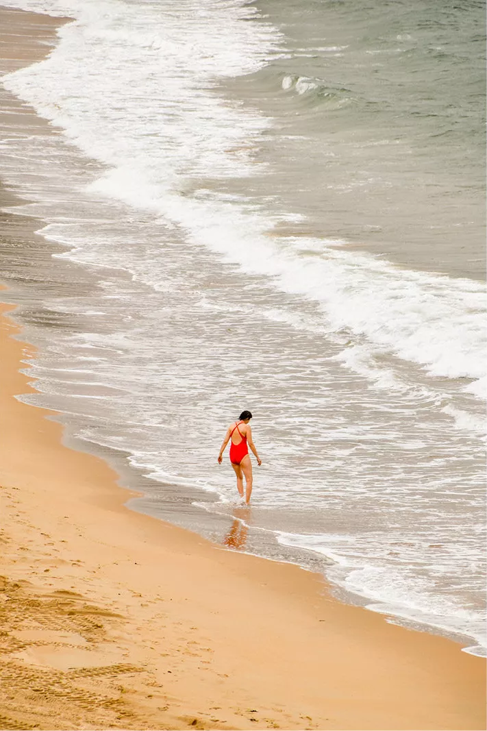 Vrouw in rood badpak wandelt de zee in, van bovenaf gezien met witte schuimlijnen op het strand
