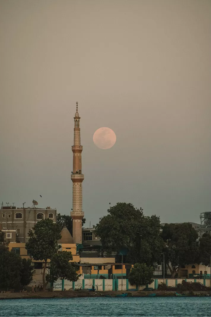 Volle maan naast de minaret van een moskee boven bomen en gebouwen aan het water