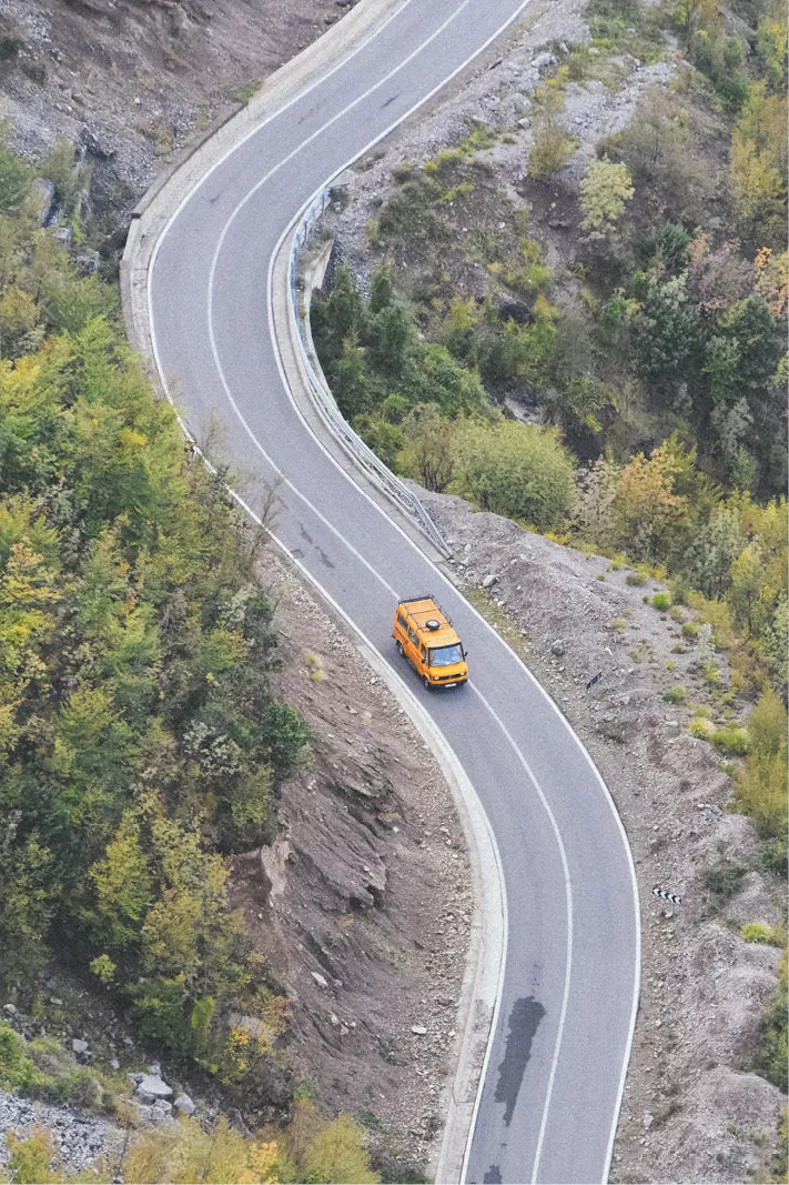 Geel busje op een kronkelende bergweg van bovenaf gezien tussen bomen en rotsen