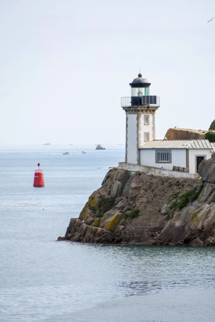 Witte vuurtoren op een rotsklif aan zee, met een rode boei in het kalme water en enkele bootjes in de verte.
