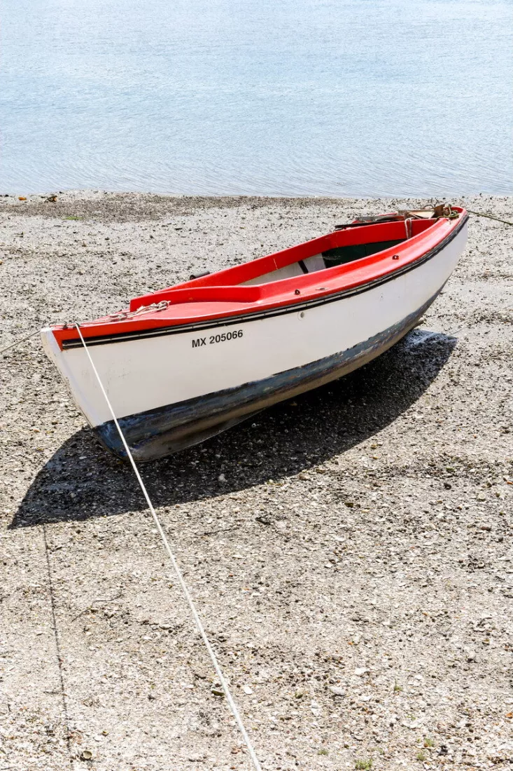 Wit roeibootje met rode rand ligt op een kiezelstrand bij laag water, met een touw naar de kant en kalm water erachter.