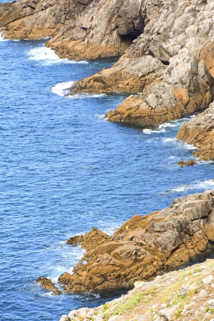 Rotsachtige kust van de Pointe du Raz met bruine rotsen, een donkere grot en blauwe zee met witte schuimkoppen.
