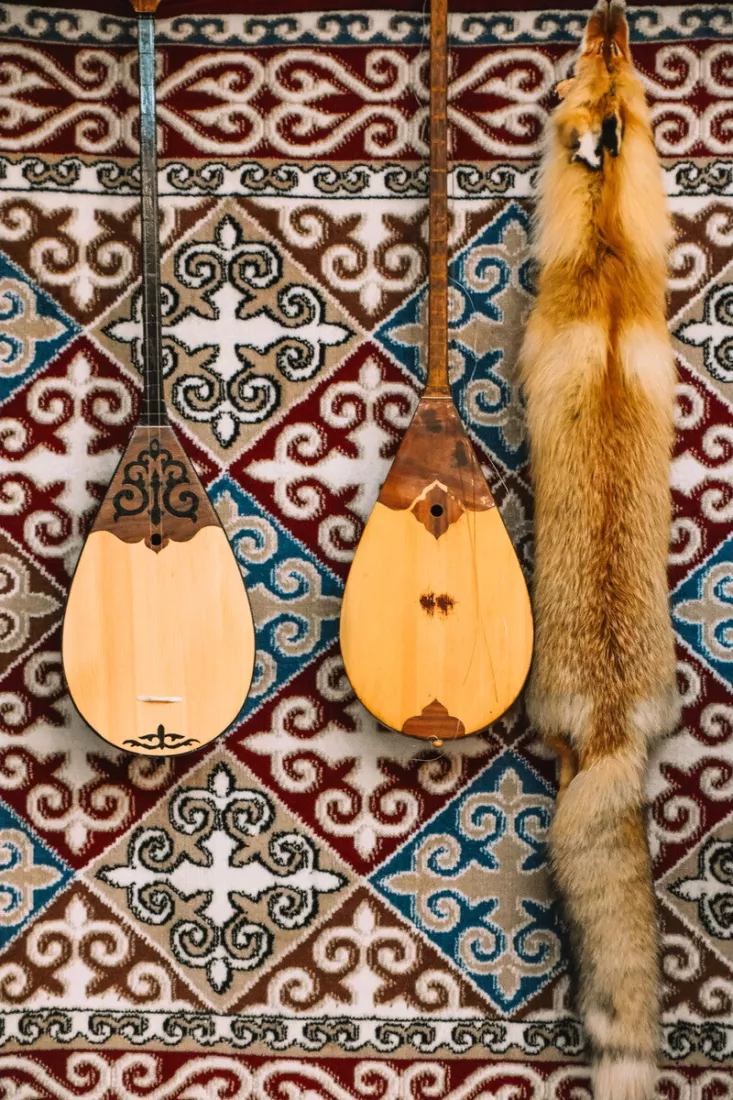 Twee dombra's en een vossenvel hangen naast elkaar voor een wandkleed met rode, blauwe en beige ornamenten.
