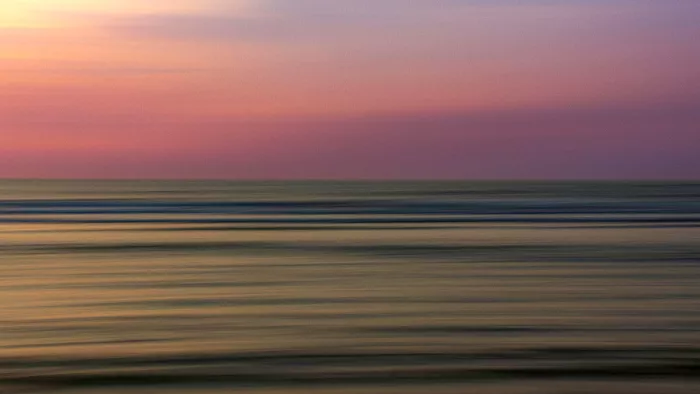 Zee en lucht in zachte horizontale banden, met een rechte horizon en kleuren van geel naar paars.
