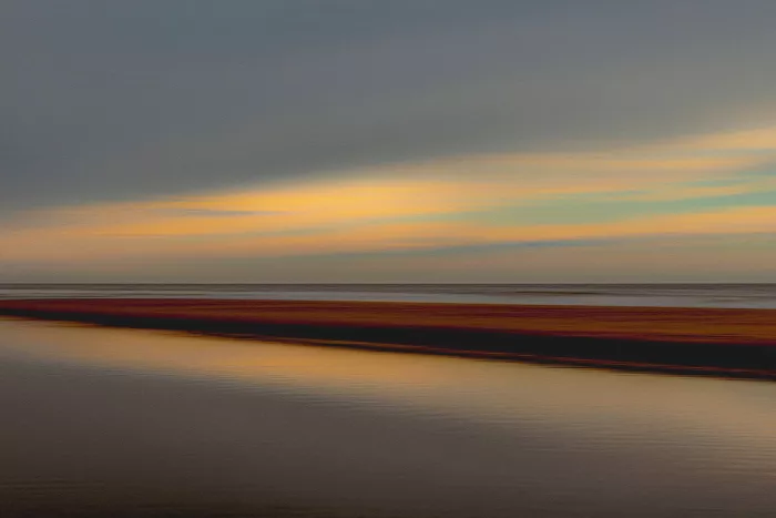 Kust met een zandbank en stil water onder een grijze lucht met gele en groenige lichtbanen boven de horizon.