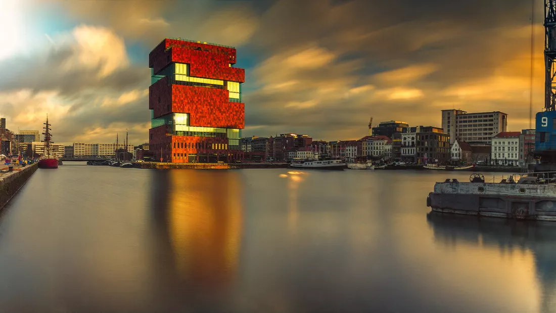 Rood torengebouw aan een havendok met uitgerekte wolken en een gouden weerspiegeling in het gladde water.