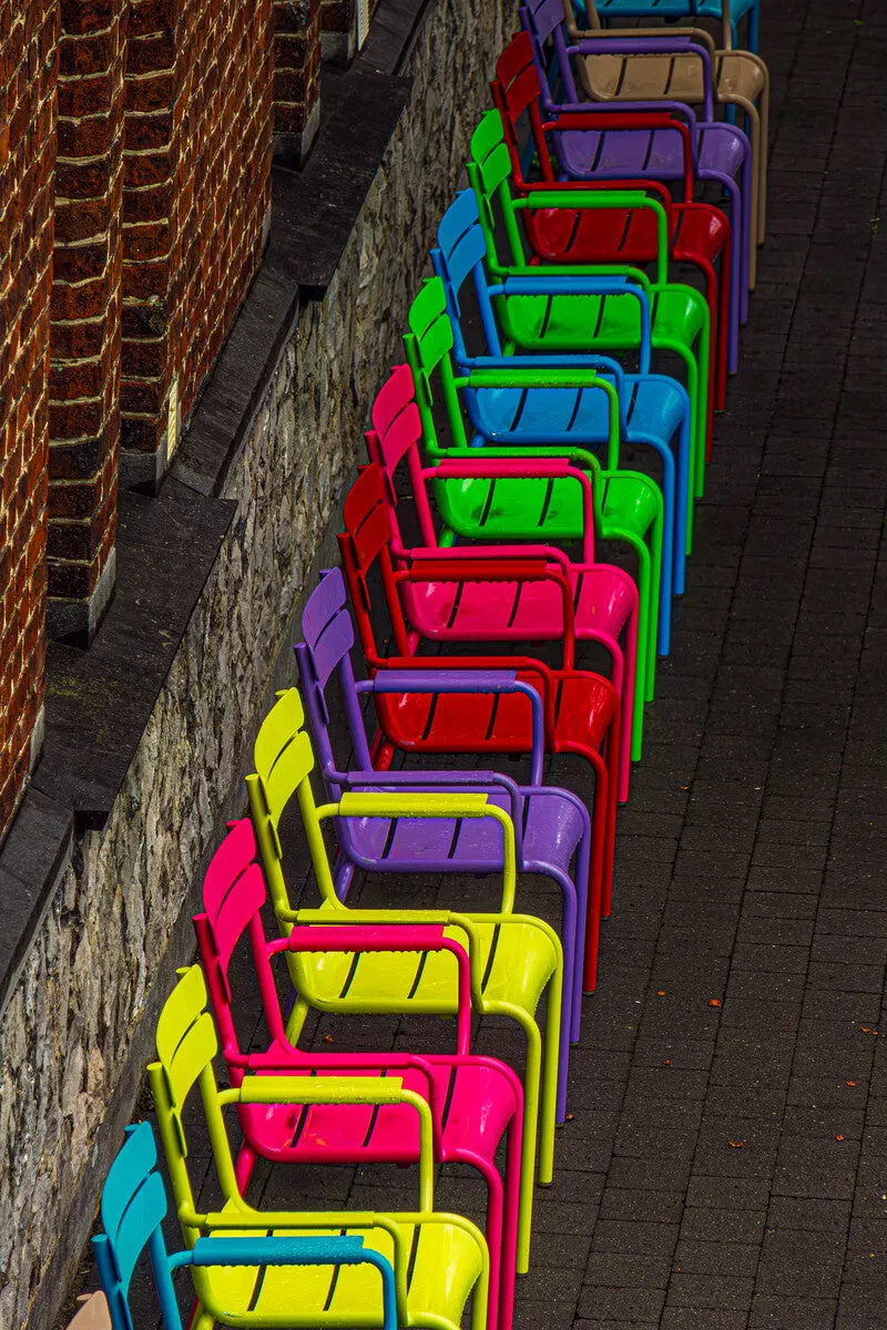 Rij gekleurde metalen stoelen in rood, blauw, groen, paars, roze en geel langs een stenen muur op donkere klinkers.