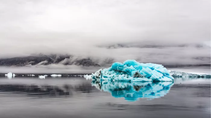 Felturquoise lage ijsberg die spiegelt in grijs water tussen mist en wolken