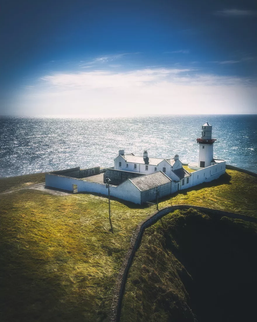 Witte vuurtoren met bijgebouwen op een grazige klif boven een glinsterende zee onder een diepblauwe lucht.