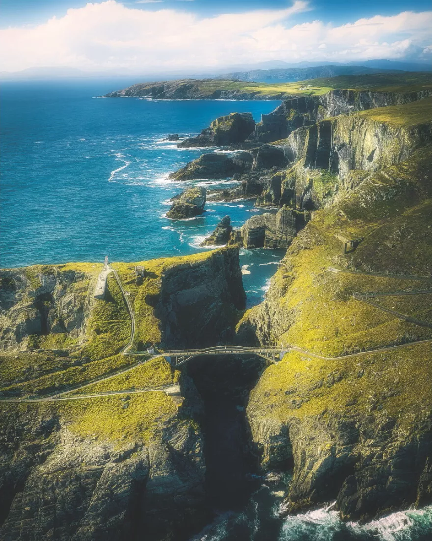 Luchtfoto van een boogbrug over een diepe kloof tussen grasgroene kliffen en blauwe zee.