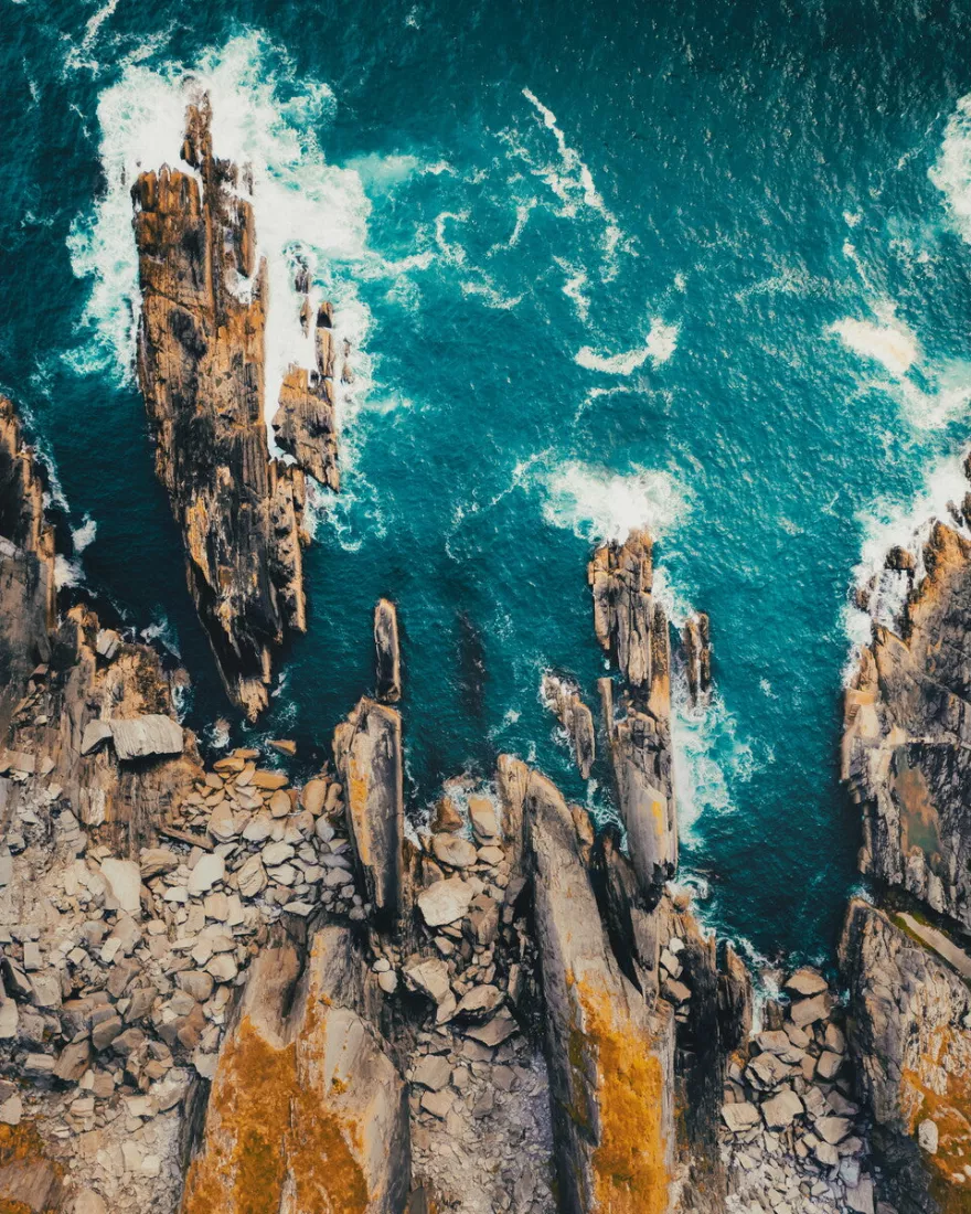 Luchtfoto van scherpe rotspunten langs een kust, met turquoise water en wit schuim van brekende golven.