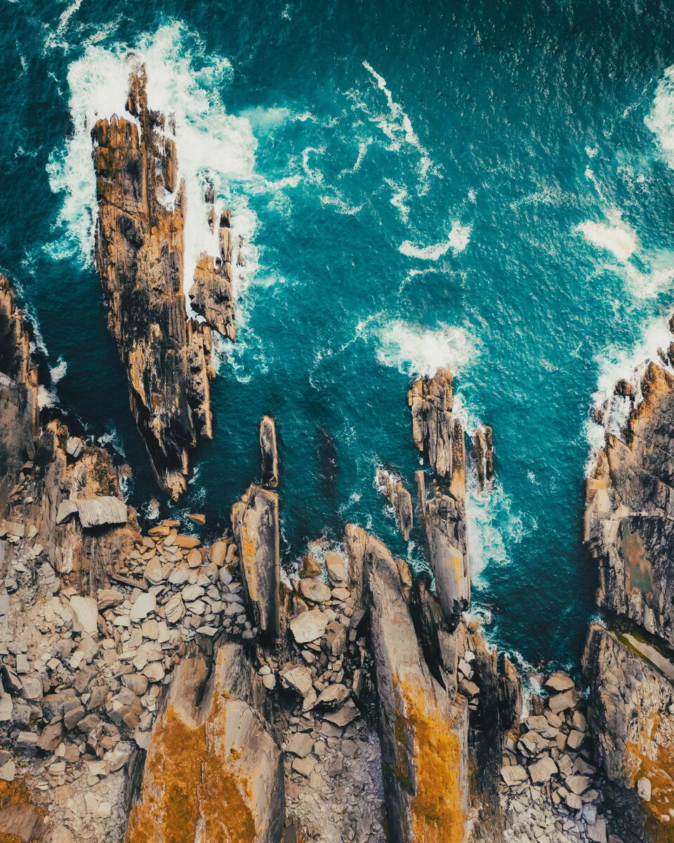 Luchtfoto van scherpe rotspunten langs een kust, met turquoise water en wit schuim van brekende golven.