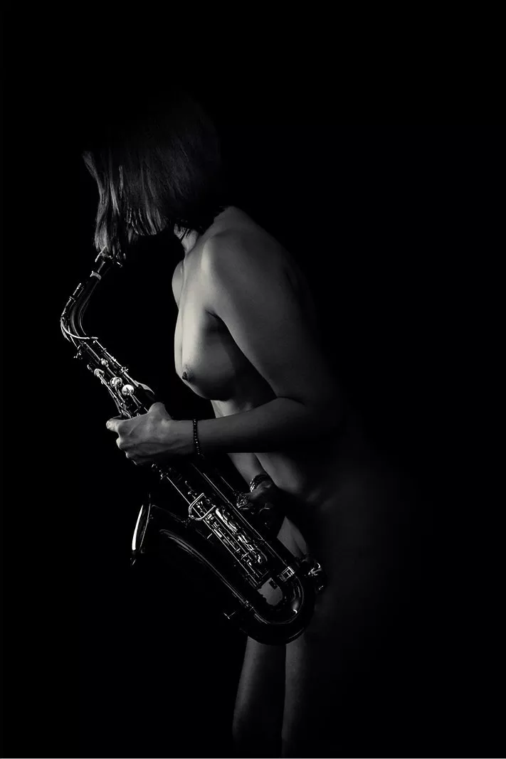 Naakte vrouw in profiel buigt over een saxofoon, met licht op het glanzende instrument in het donker