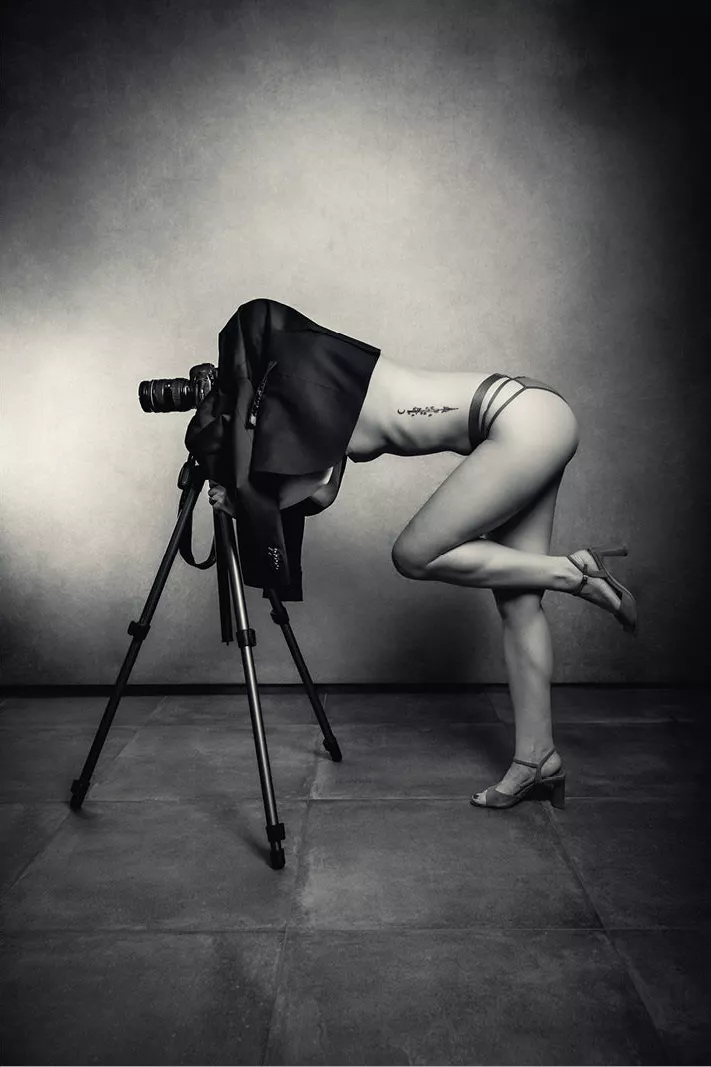 Vrouw met het hoofd onder een donkere doek achter een camera op statief, één been omhoog, in een grijze studio