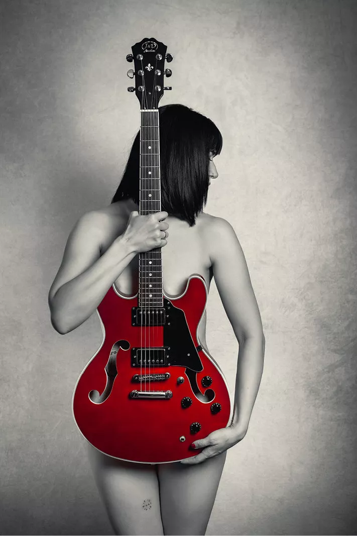 Naakte vrouw achter een rode gitaar met de hals voor het gezicht, zwart-wit met rode gitaar
