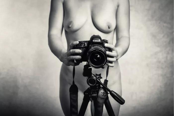 Naakte vrouw achter een camera op statief met de lens naar de kijker, uitgesneden in breed formaat