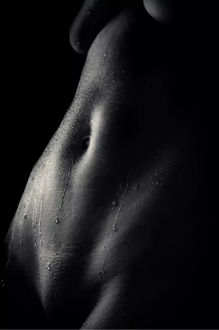 Close-up van een vrouwelijke buik met navel en waterdruppels in strijklicht, low-key zwart-wit