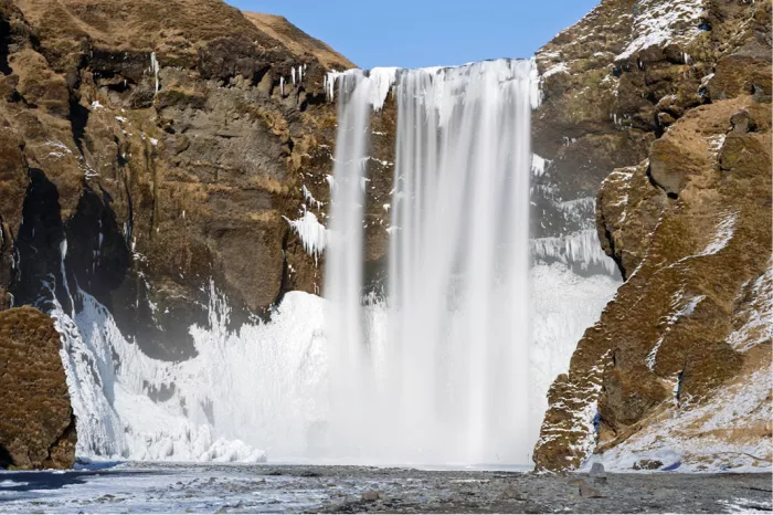 Brede waterval tussen bruine rotswanden met ijs, sneeuw en ijspegels aan de voet