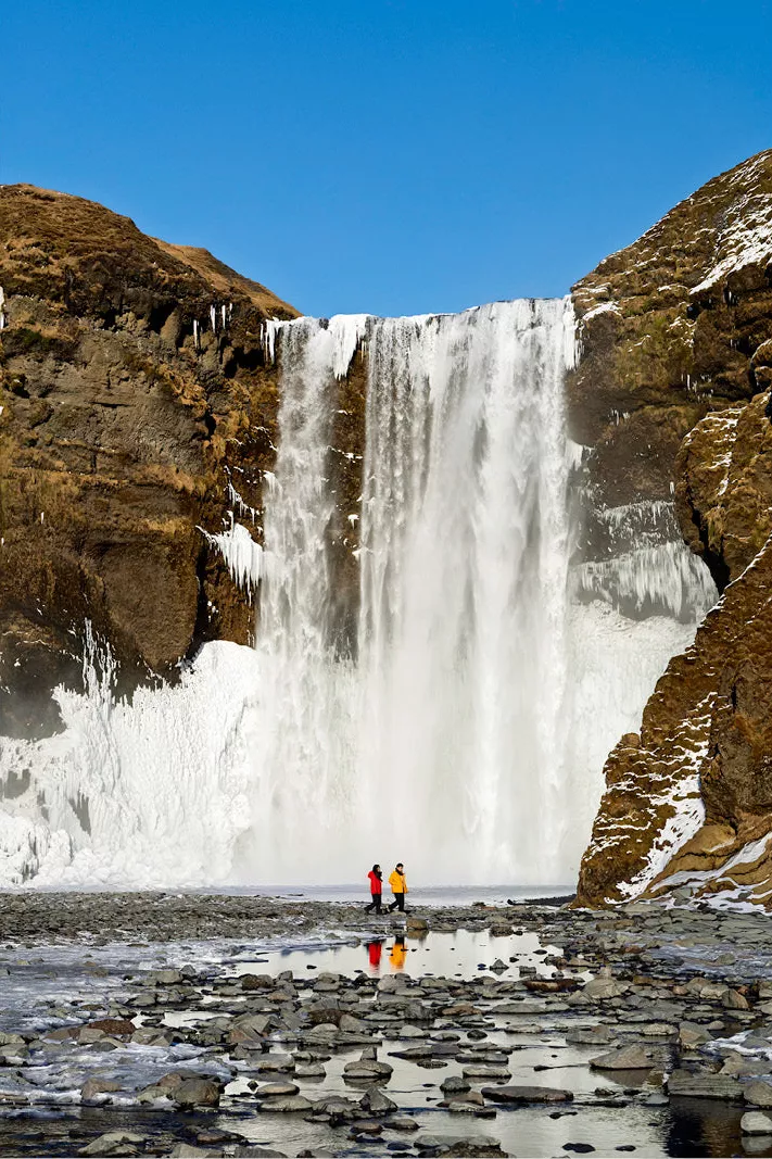 Hoge waterval met ijs, met twee kleine figuren in rood en geel die spiegelen in het water