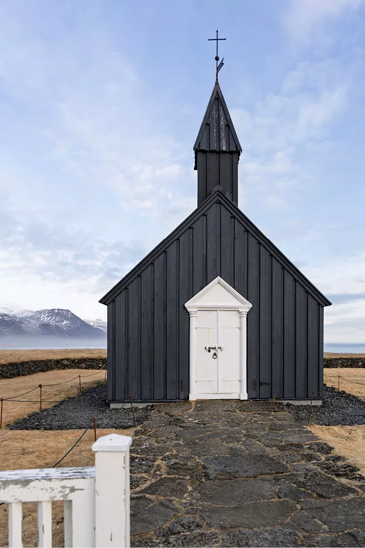 Zwarte houten kerk met spitse toren en witte deur aan het einde van een stenen pad