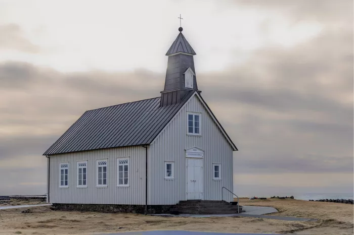 Wit houten kerkje met grijs dak en slanke toren bij de zee onder een grijze lucht