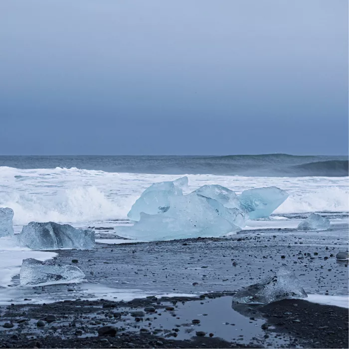 Blauwe ijsblokken op een zwart strand met aanrollende witte golven onder een grijze lucht