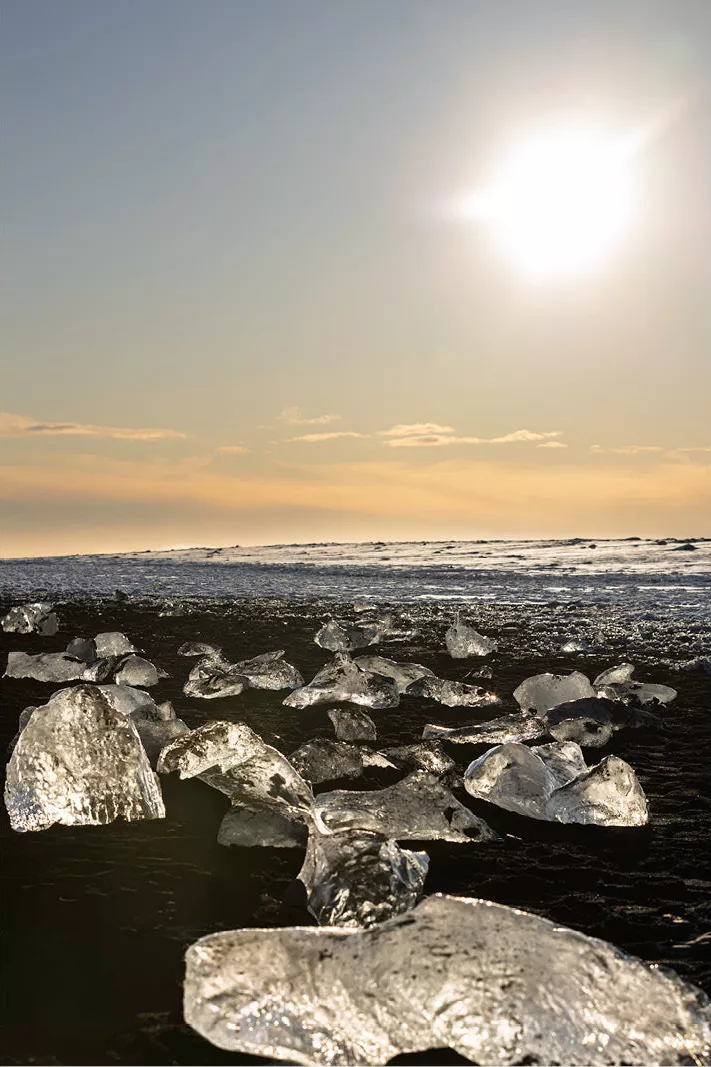 Felle zon boven een zwart strand met verspreide, glanzende ijsblokken in tegenlicht