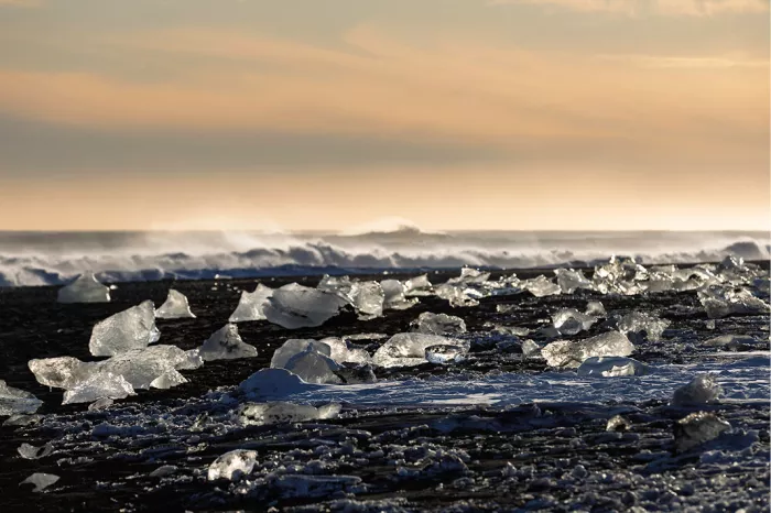Glinsterende ijsblokken op een zwart strand met brekende golven onder een oranje lucht
