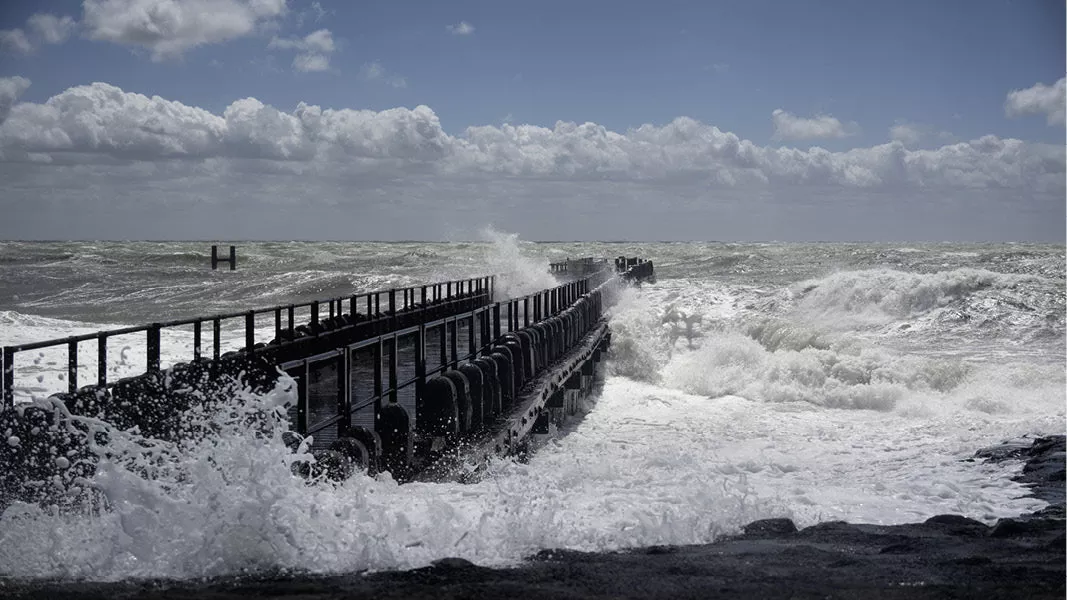 Witte golven die tegen een donkere houten pier slaan onder een blauwe lucht met stapelwolken
